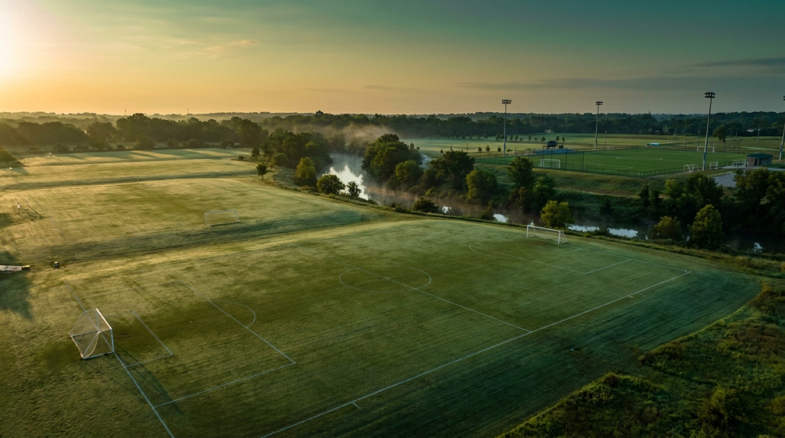 An aerial view of a soccer complex at sunrise, with several grass fields, mist over the creek and a lit turf field in the distance